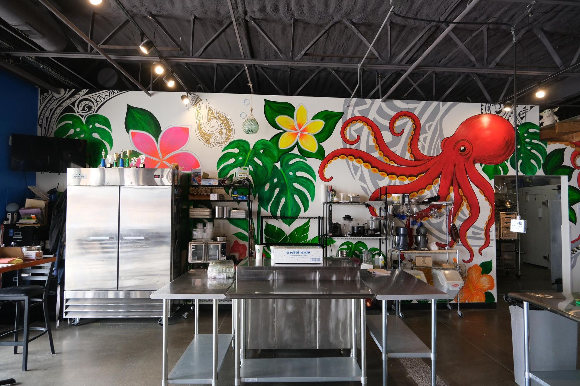 A mural of a red octopus, tropical flowers, and green leaves on a wall behind stainless steel kitchen equipment.