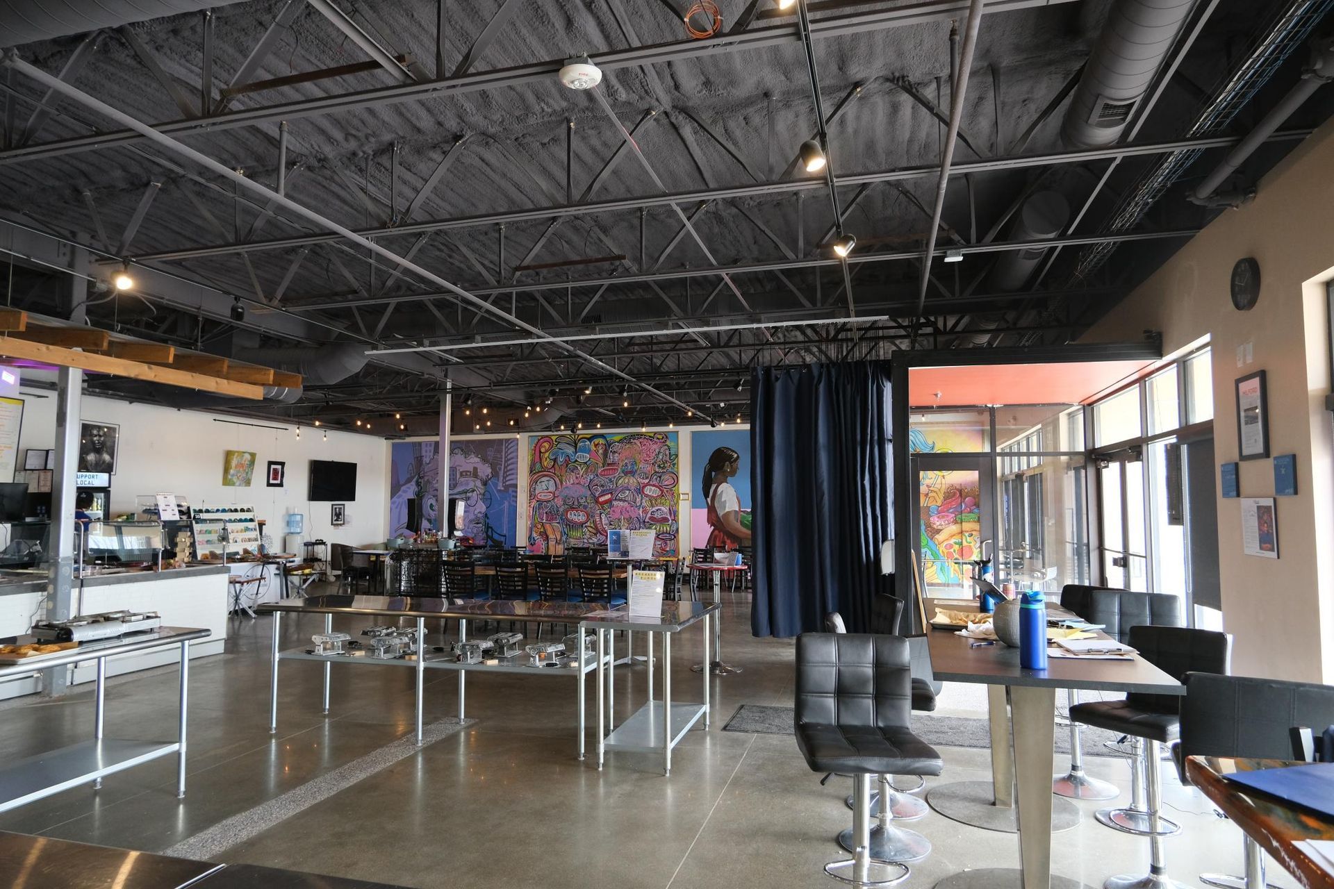 Spacious cafe interior with high ceilings, exposed trusses, modern furniture, and large colorful murals on the walls.