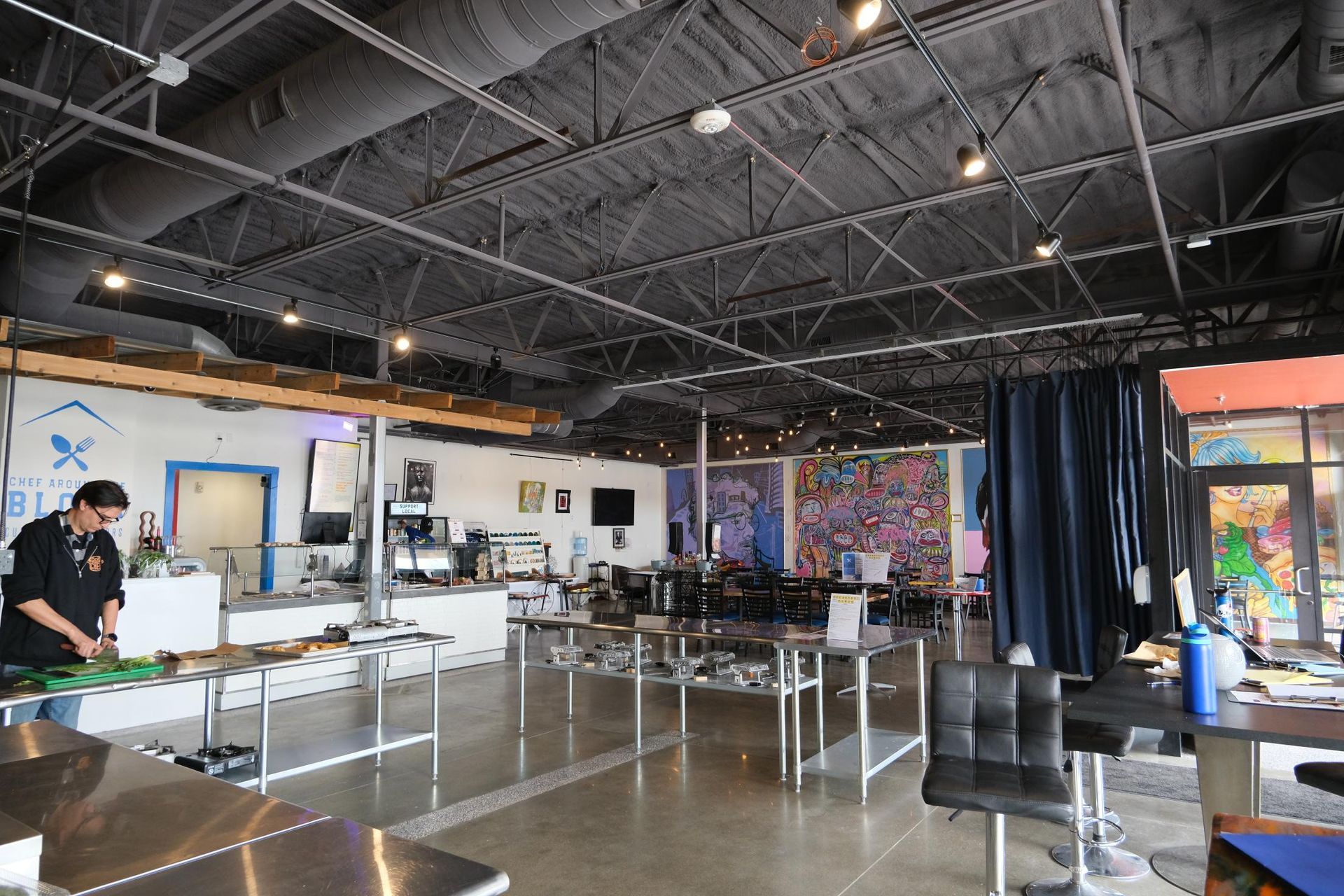 A person prepares food in a kitchen area with an open-concept dining room, high industrial ceilings, and colorful art.