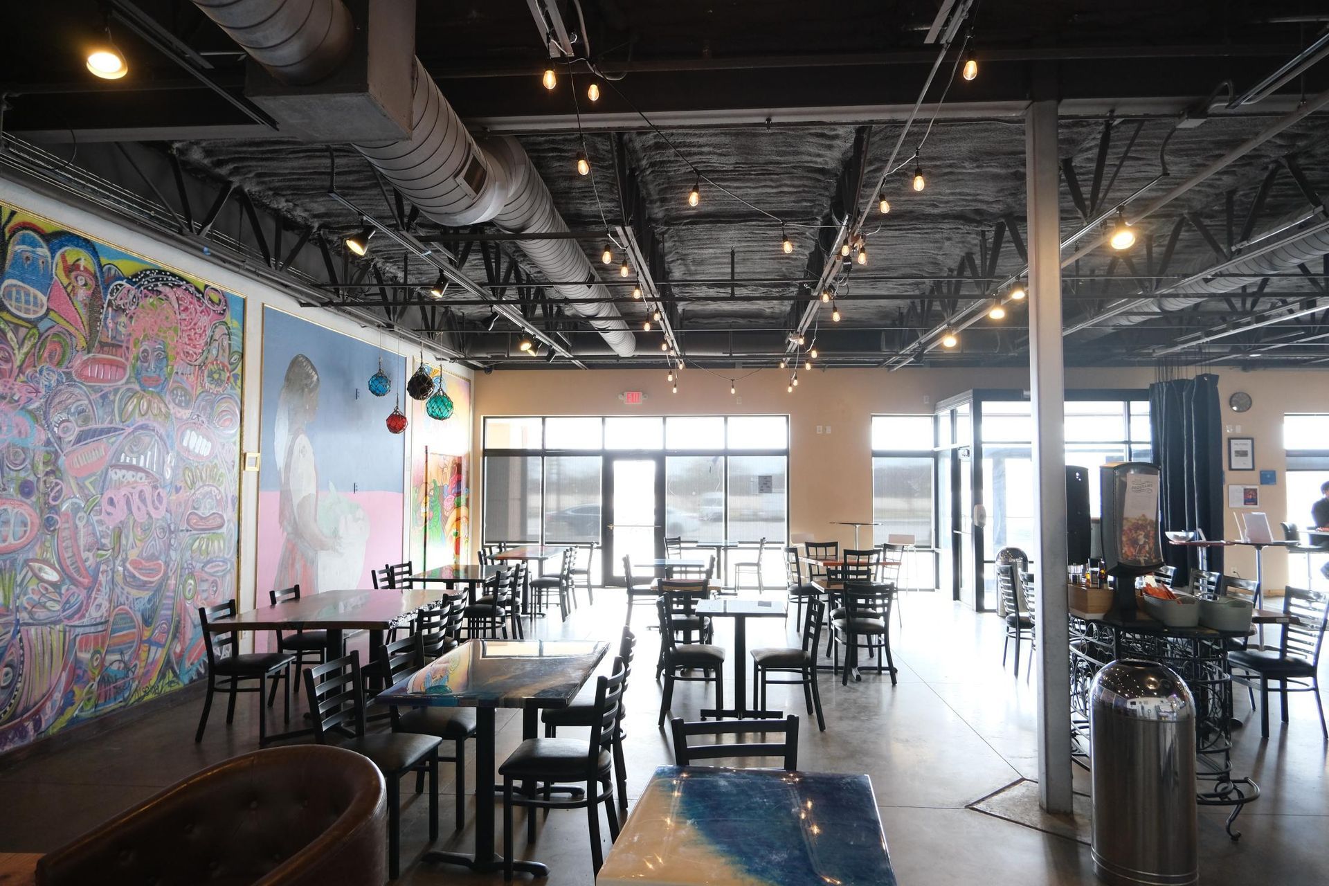 An indoor cafe with tables and chairs, a vibrant mural on the left wall, and an exposed ceiling with industrial lighting.