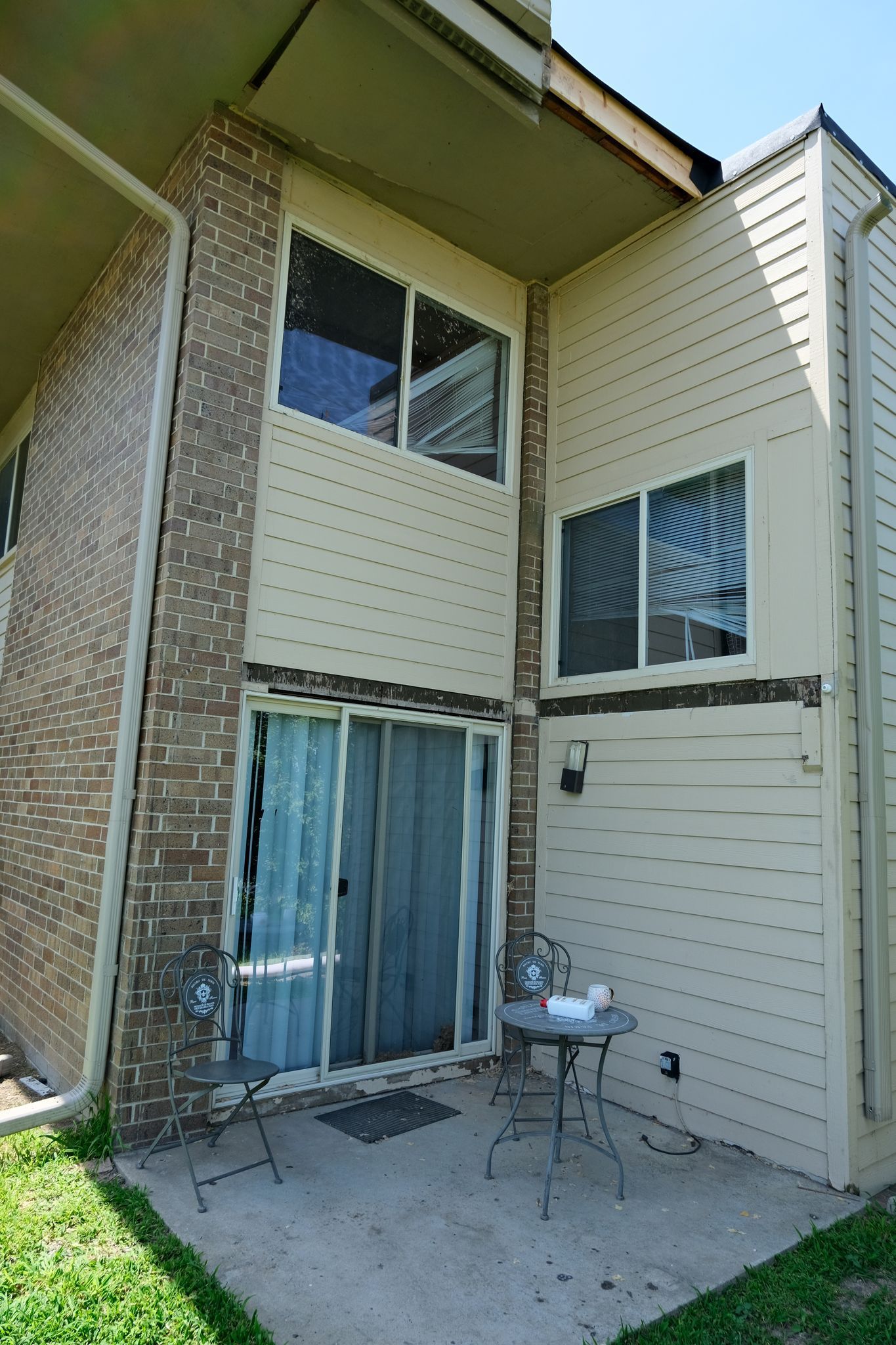 Exterior of a two-story building with tan siding, a brick column, sliding glass doors, and a small patio.