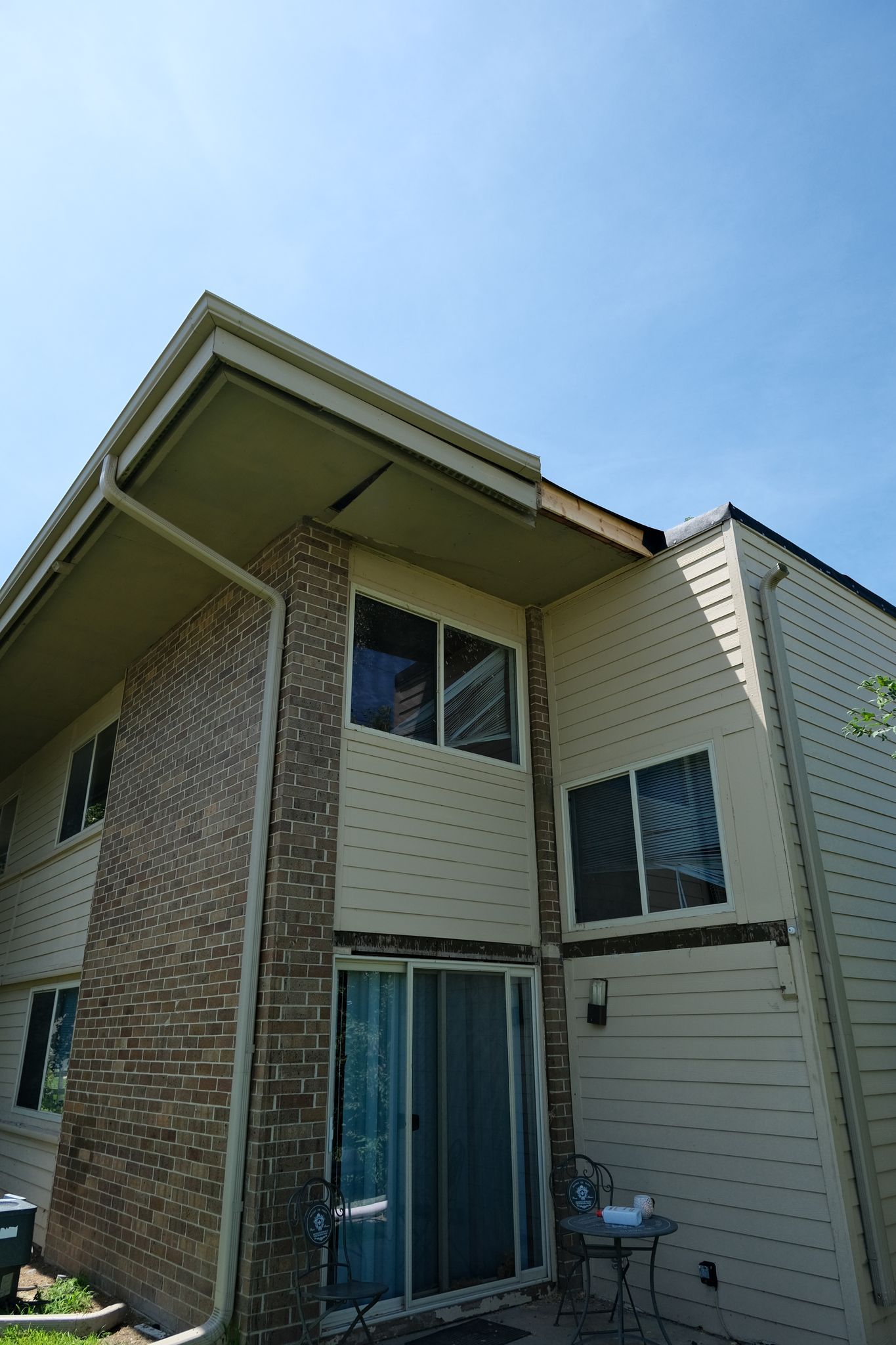 Two-story building exterior with beige siding, brick chimney, windows, and sliding glass door against a blue sky.