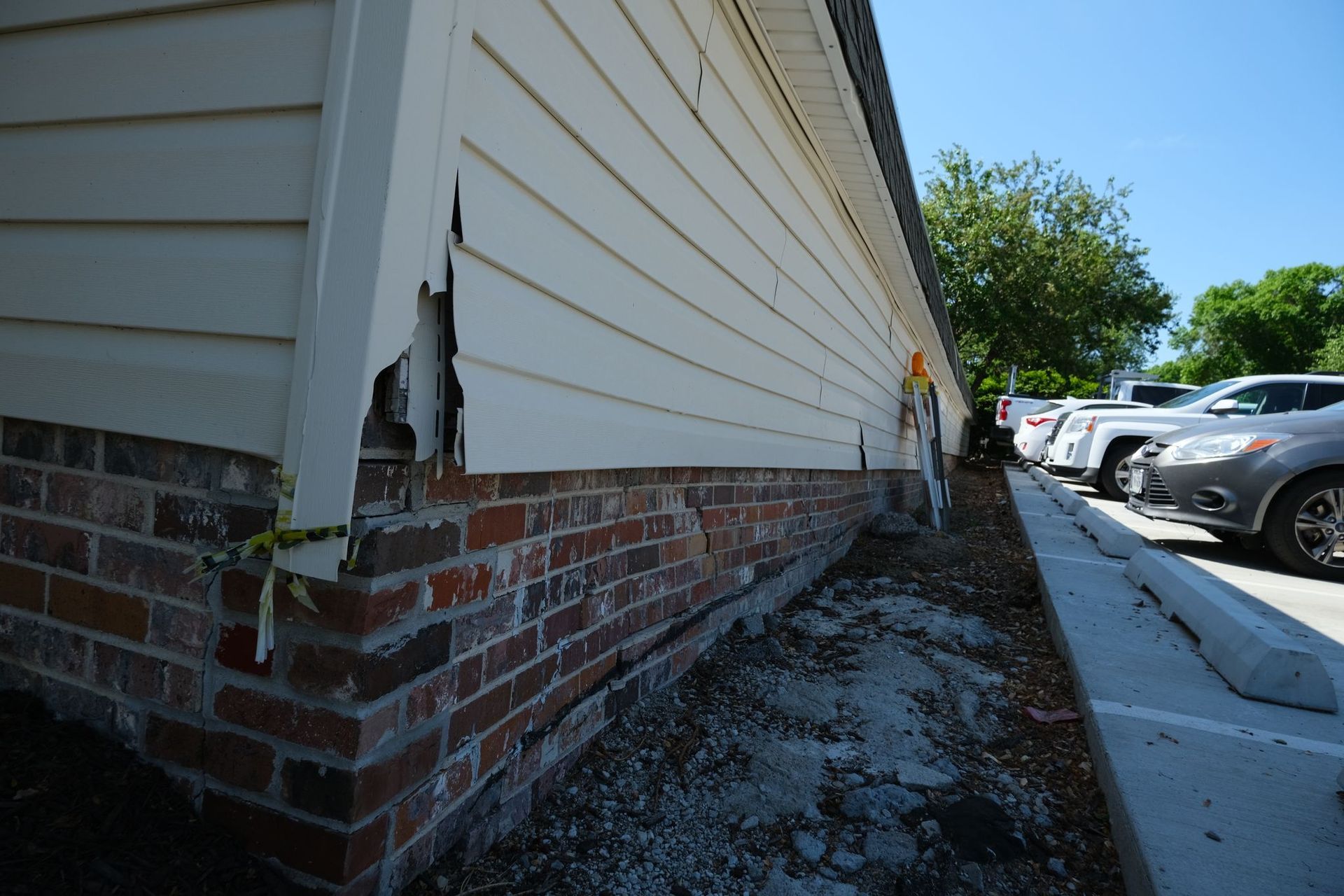 Damaged building exterior: brick base, white siding with tear, parked cars and trees in background.
