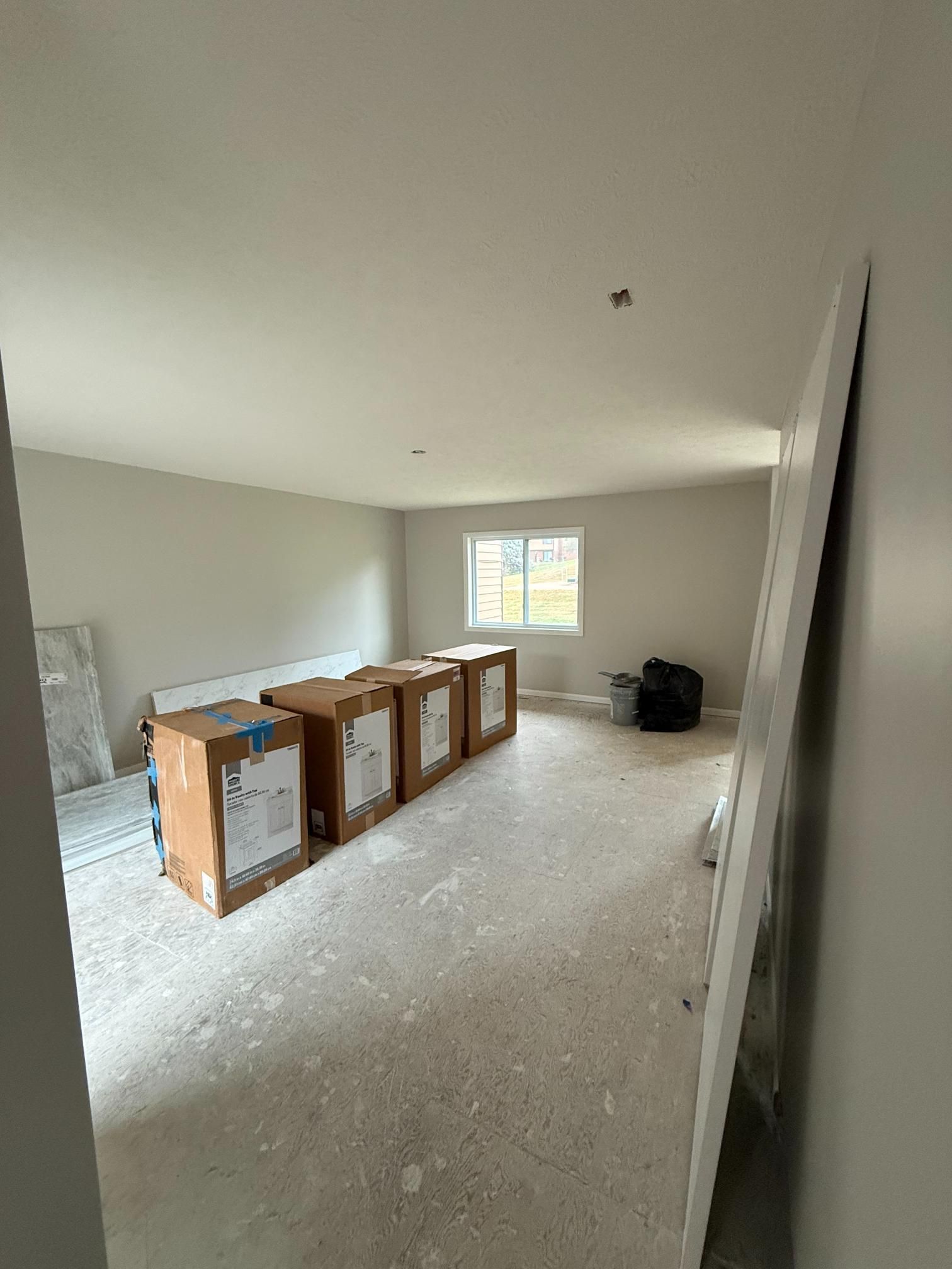 Interior of a room under construction; cardboard boxes in the center, window in the back, bare floor, and off-white walls.