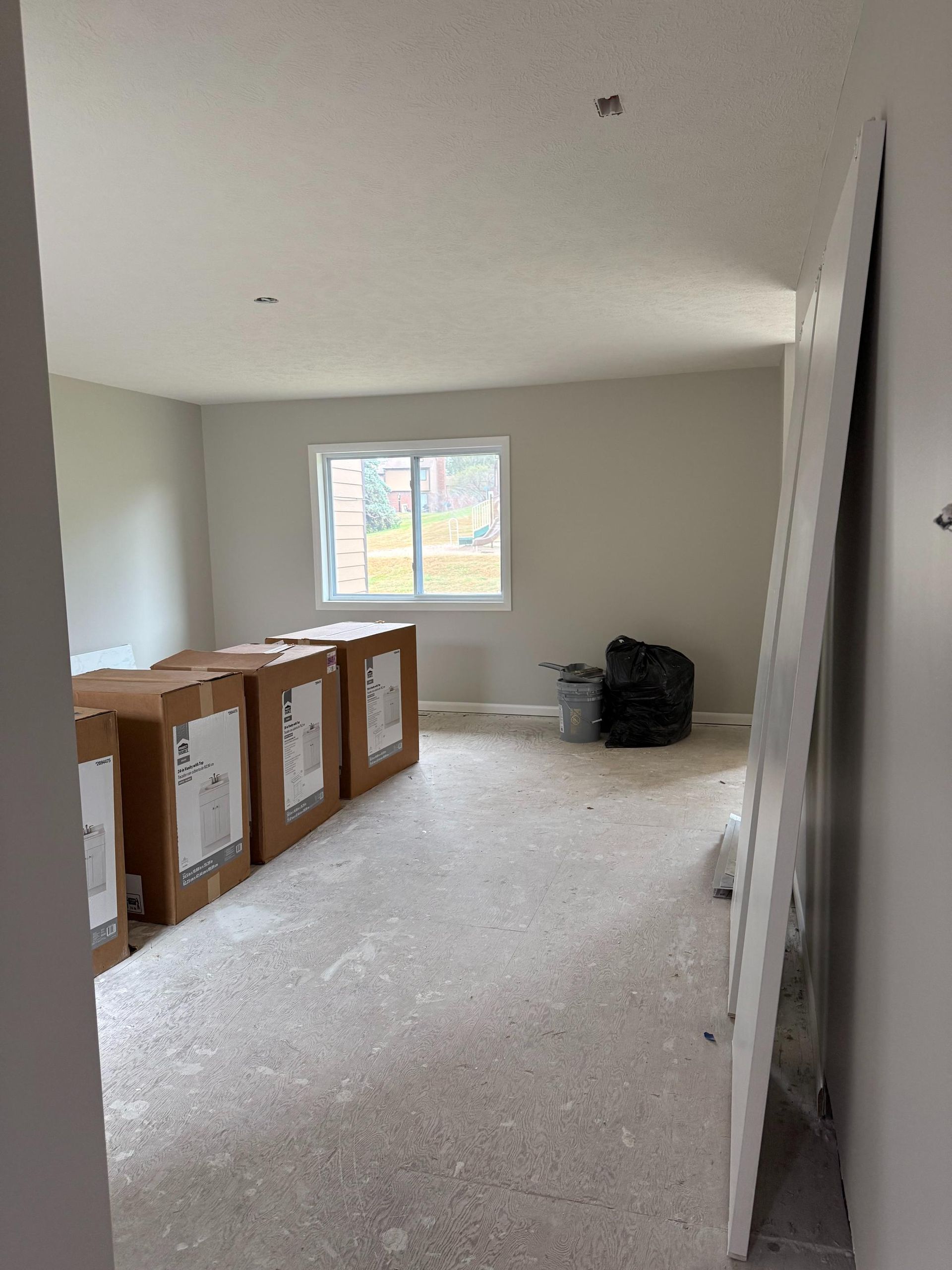 Interior of a room with unfinished flooring, window, and cardboard boxes. A sheet of drywall leans against the doorframe.