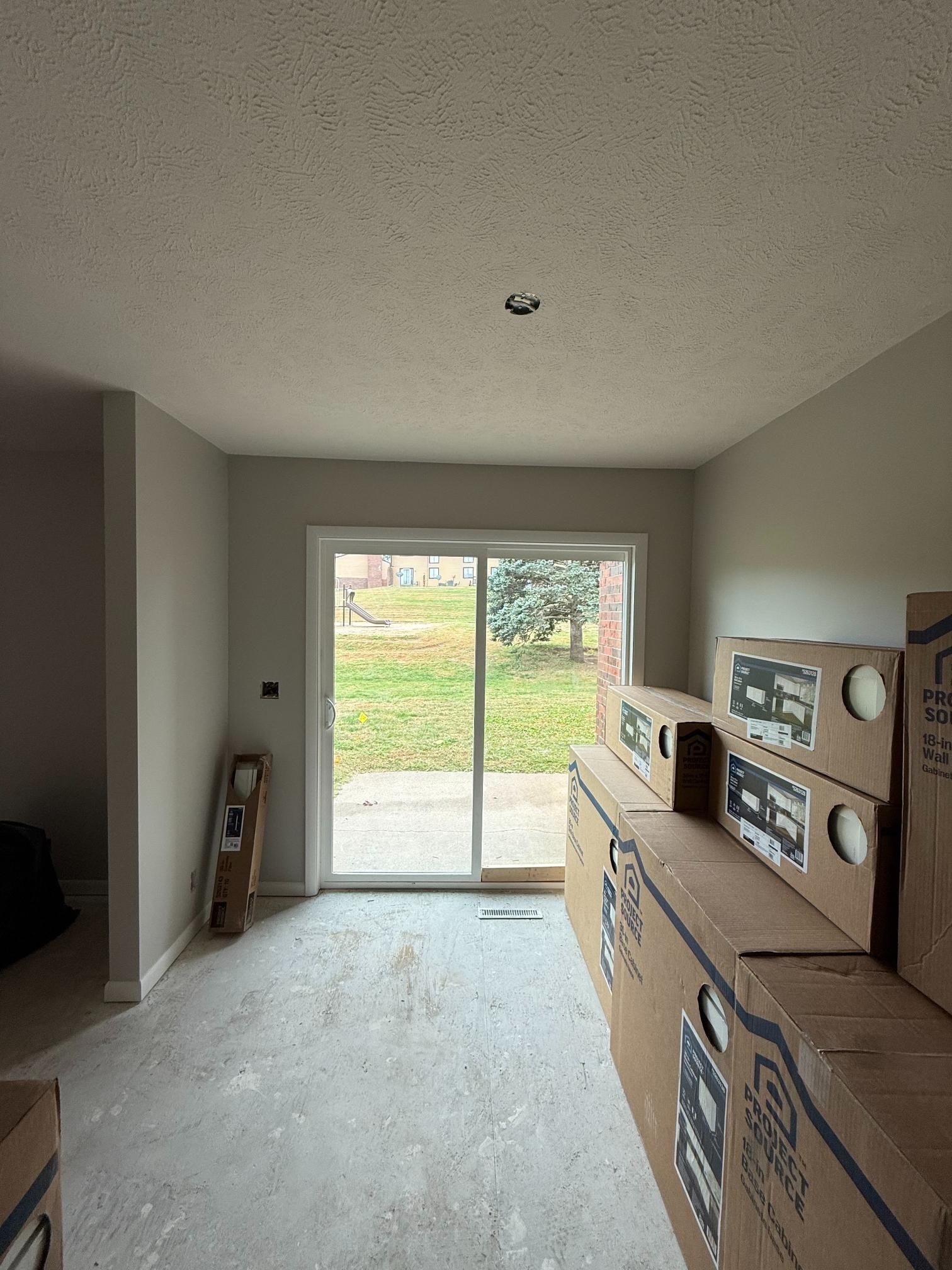 Room with sliding glass door, beige walls, stacks of cardboard boxes. Grass and trees visible outside.