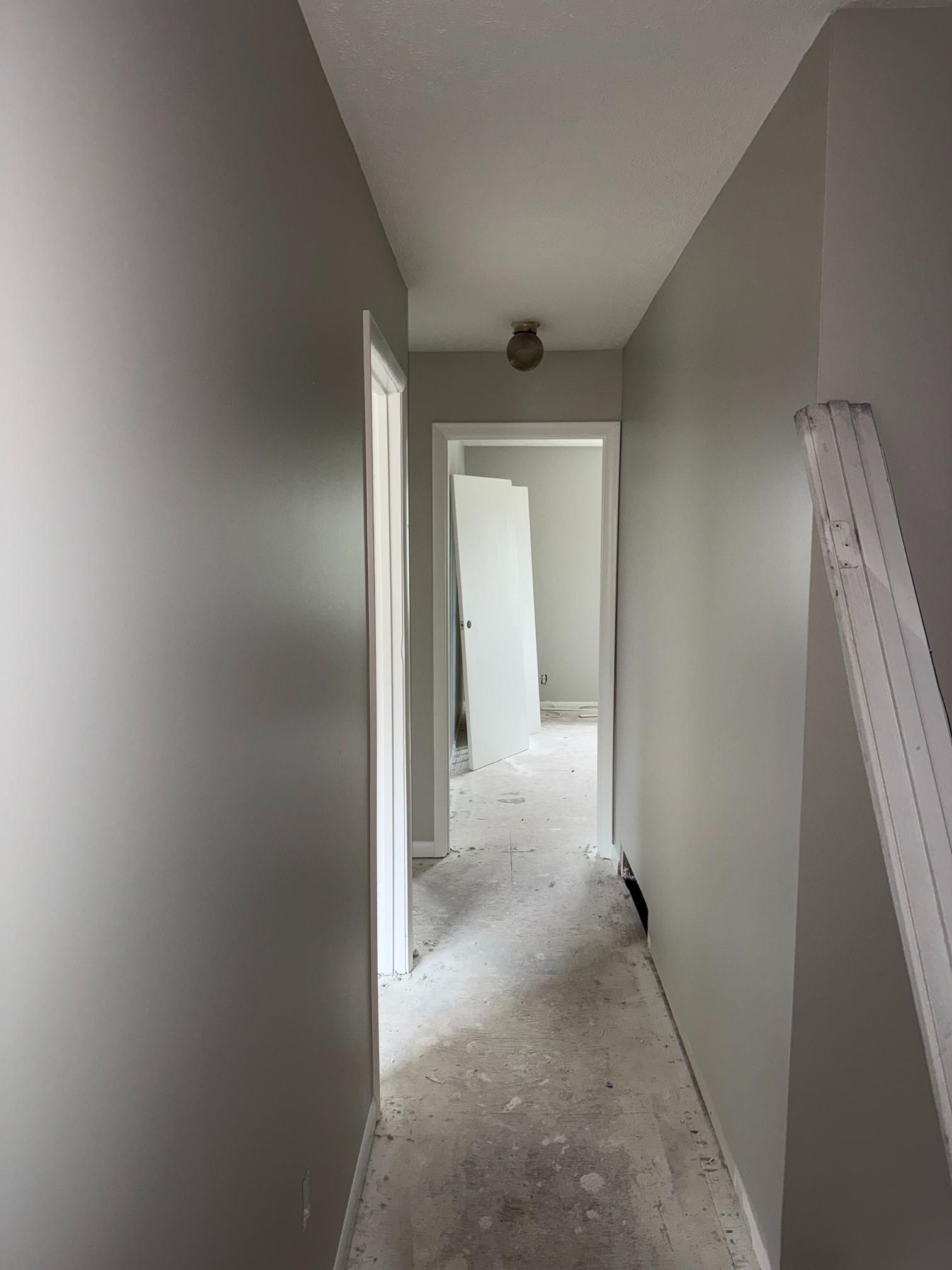 Narrow hallway with gray walls, unfinished floor, open doors, and ceiling light.