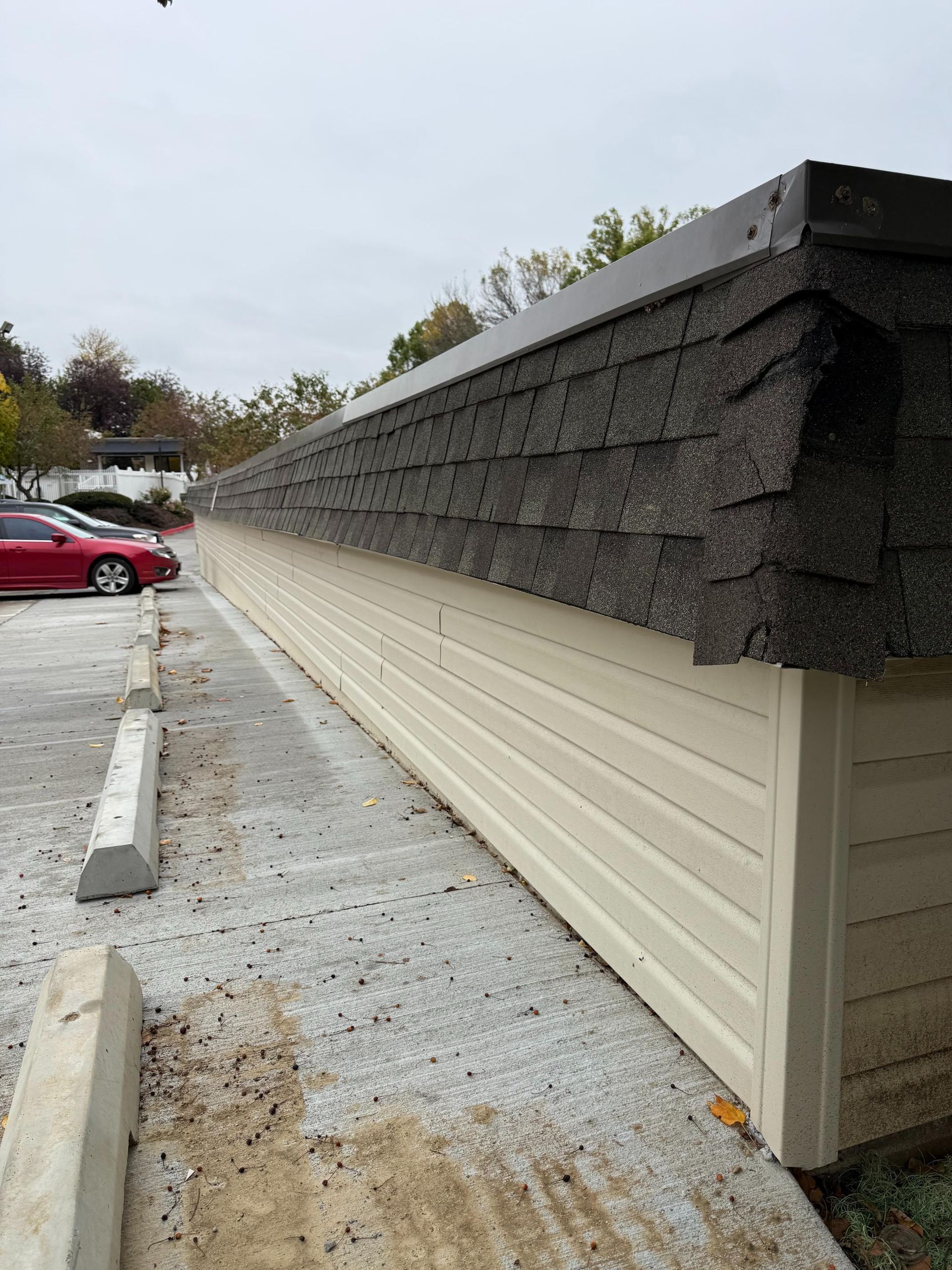 Long, light-colored building with dark roof in parking lot with car, concrete barriers, and gravel.