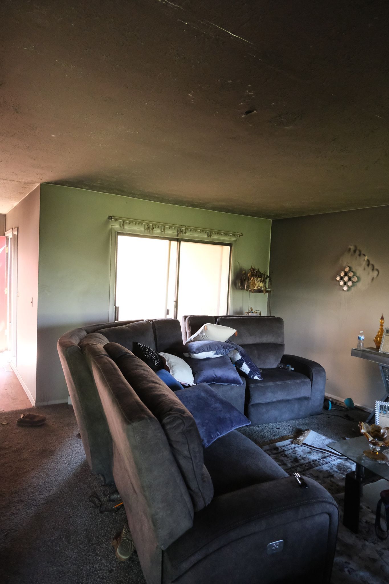 Living room with dark recliner sofa, window, and grey walls. Ceiling shows damage.