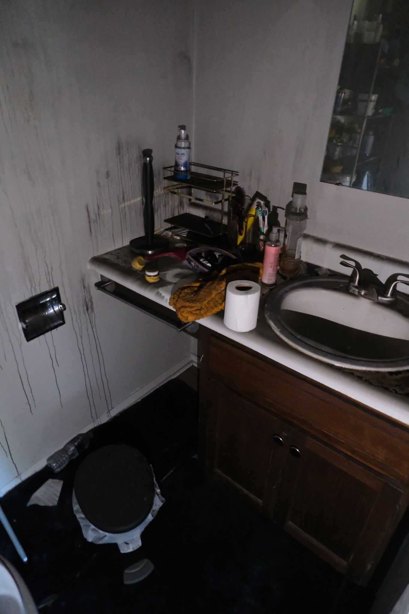 Bathroom with a cluttered vanity, toilet, and mirror. The walls are stained.