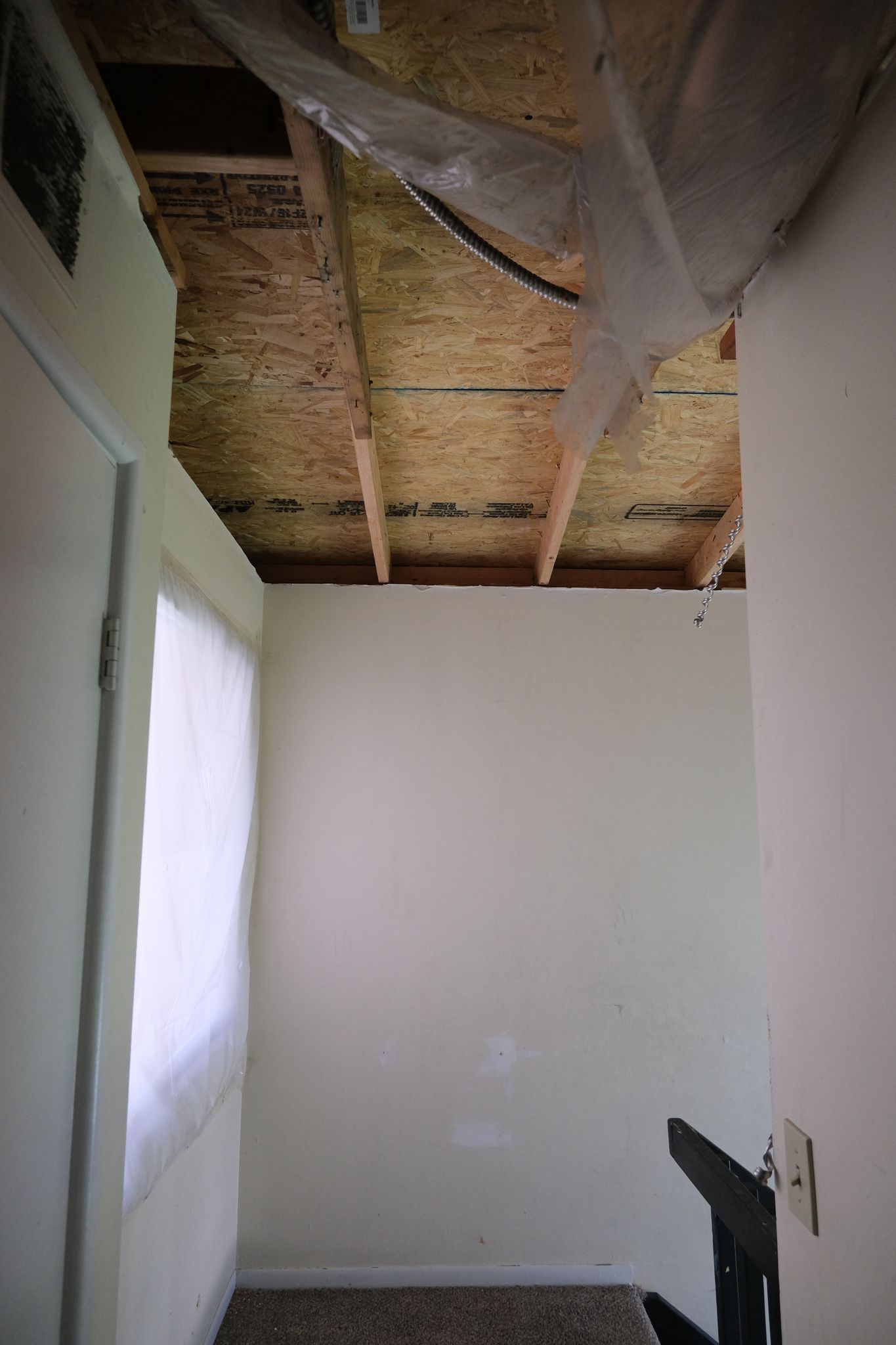 Interior view of a room with a partial ceiling. White walls, window with white curtain, and a dark stairway are visible.