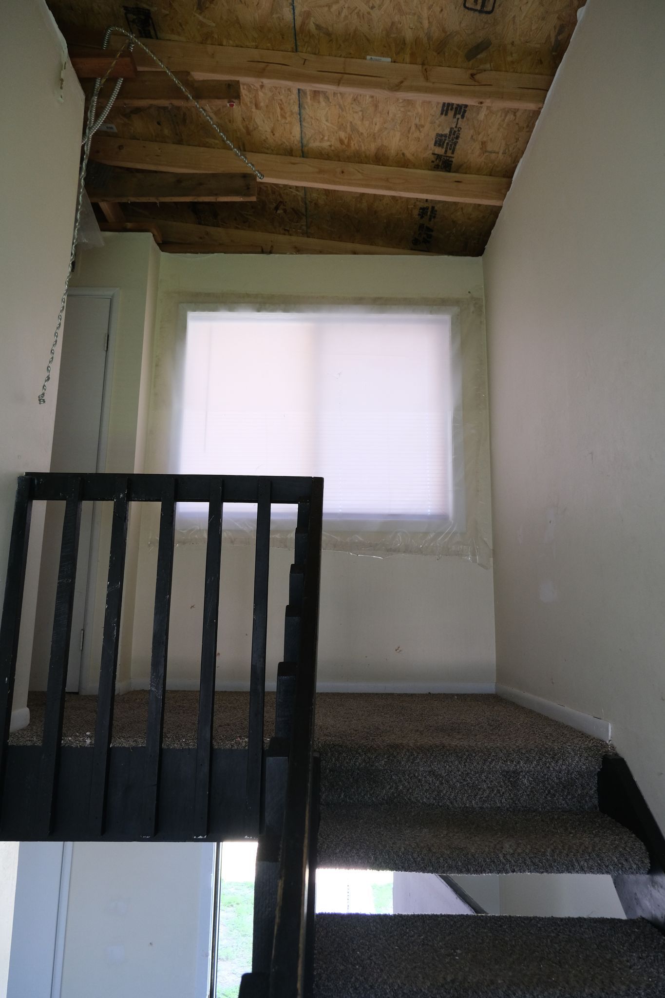 Staircase leading up to a window with white shades. Dark railing on left. The ceiling is unfinished.