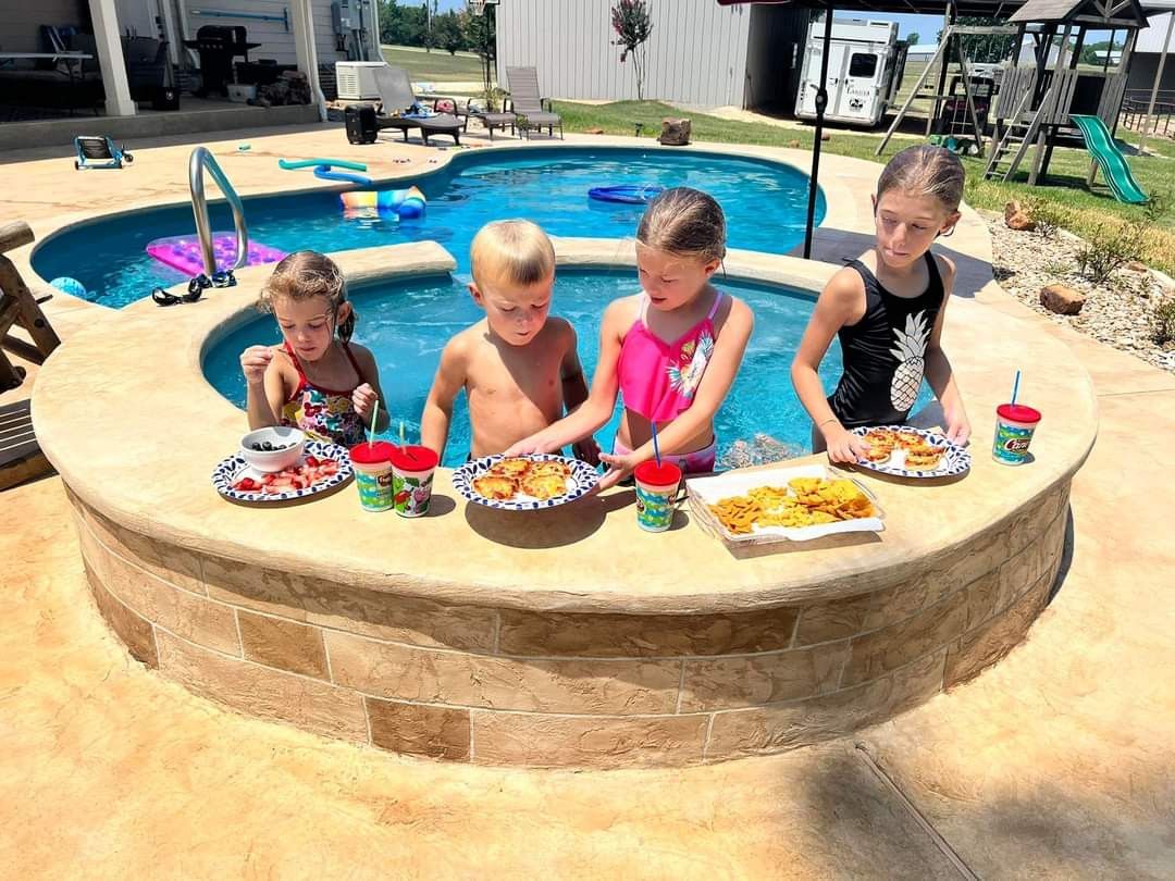 A group of children are sitting at a table in front of a swimming pool.