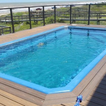 A large swimming pool is sitting on top of a wooden deck next to the ocean.