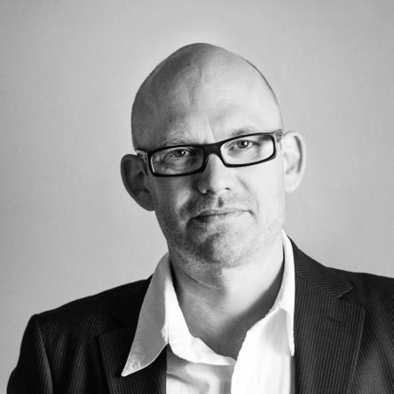 Auf einem Schwarzweißfoto blickt ein kahlköpfiger Mann mit Brille und Anzug in die Kamera.