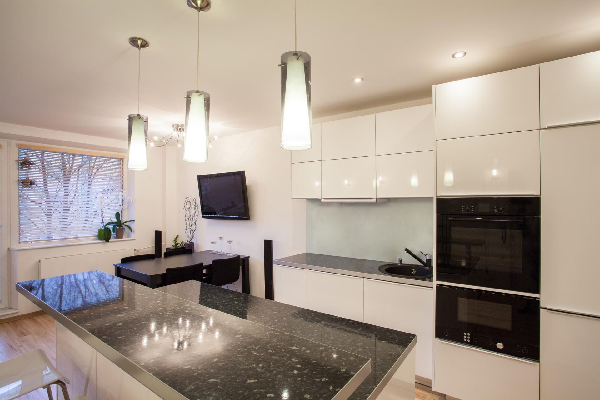 Modern white kitchen with black granite island, pendant lights, built-in oven, and wall-mounted TV.