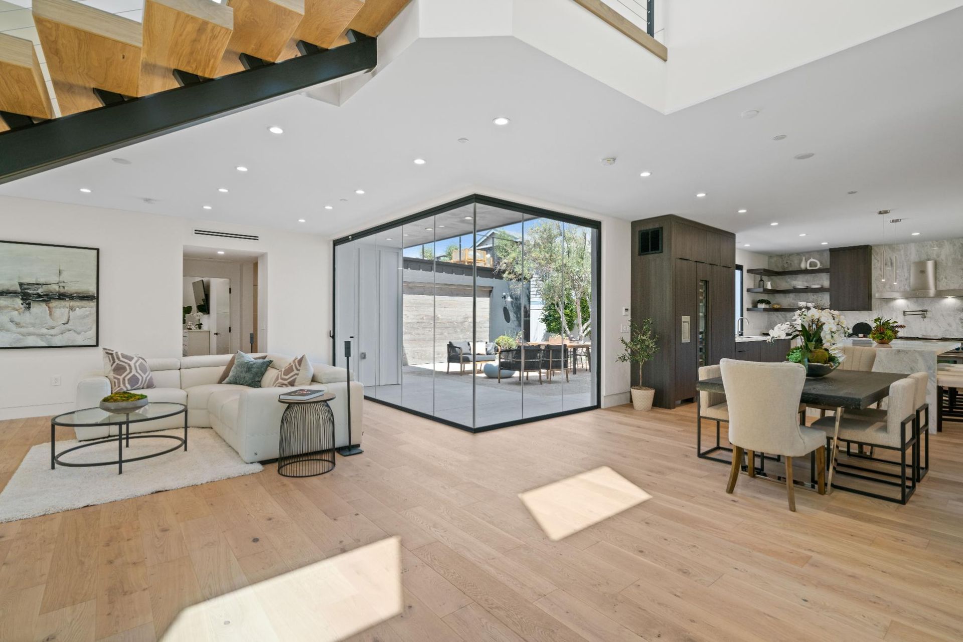 Modern open-plan living room and dining area with light wood floors, white furniture, and large glass doors to a patio