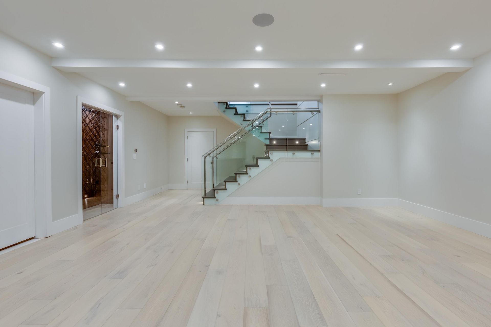 Empty modern living room with light wood floors, recessed lights, and glass staircase railing