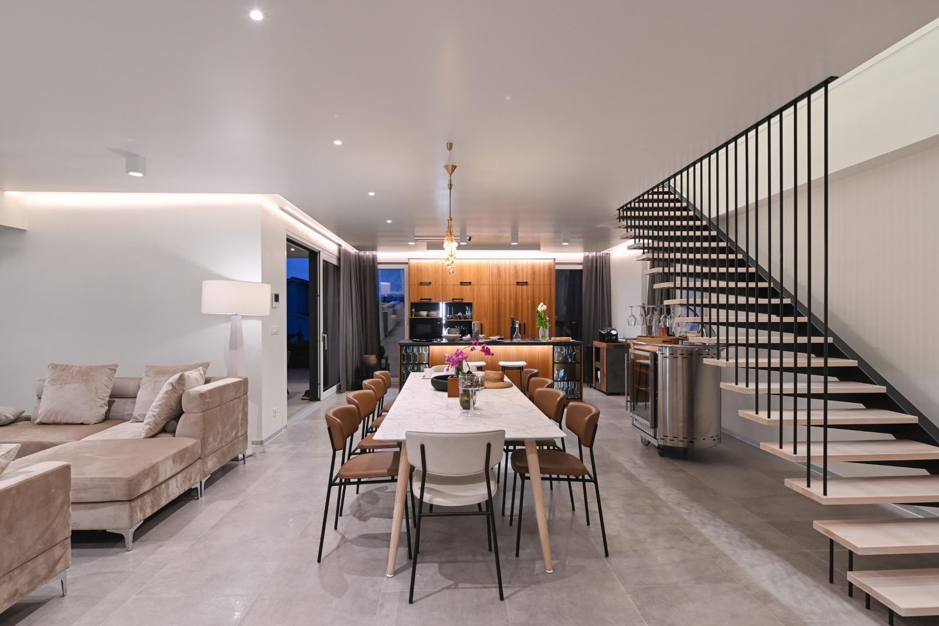 Modern open-concept living and dining room with a staircase, neutral decor, and warm kitchen lighting
