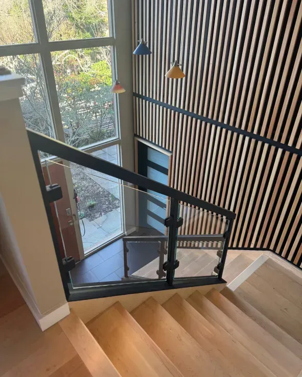 Staircase with wood steps and black railing, glass panel, wood slat wall, large window with view.