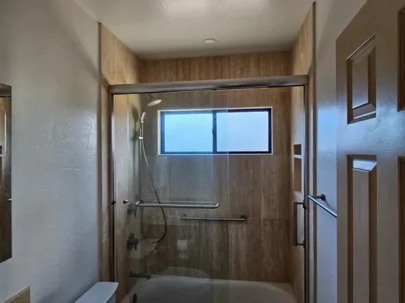 Bathroom with shower, tub, and window; neutral-toned wall tiles, glass door, and silver grab bars.
