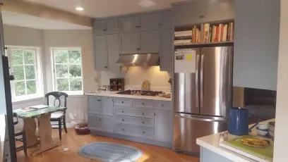 Kitchen with blue cabinets, stainless steel refrigerator, and windows; a small table and chair sit in front of the windows.