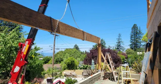 Crane lifts wooden beam during construction outdoors on a sunny day.