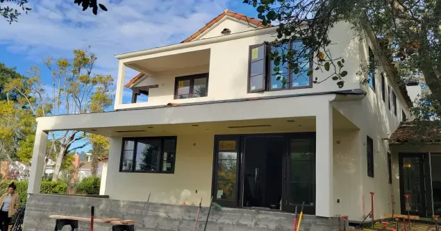 Two-story cream-colored house with large windows, covered porch, and partial landscaping under a blue sky.