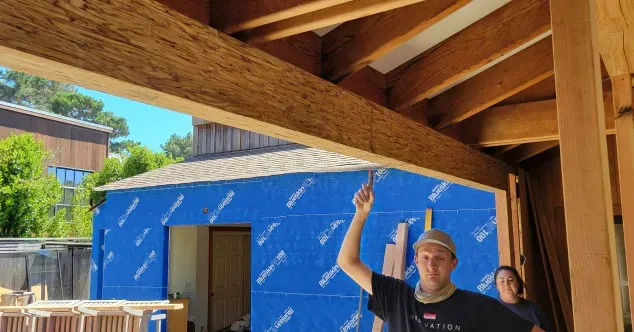 A person points up at a wooden beam, construction site. Building with blue insulation and a person standing nearby.