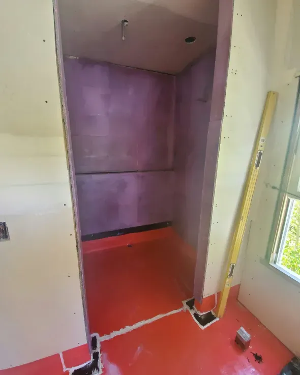 Red waterproof membrane installed in a small shower enclosure, with purple walls. A level leans against the side.