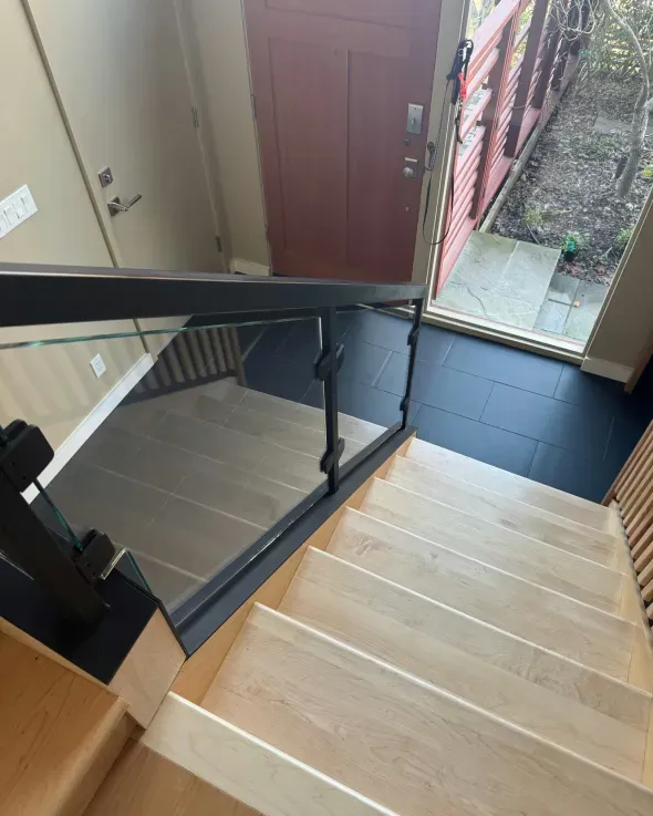 Staircase with light wood steps, glass and black metal railing, leading to a dark floor near a red door.