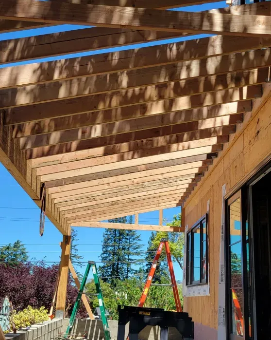 Wooden pergola under construction, attached to a house. Ladders and tools are present.