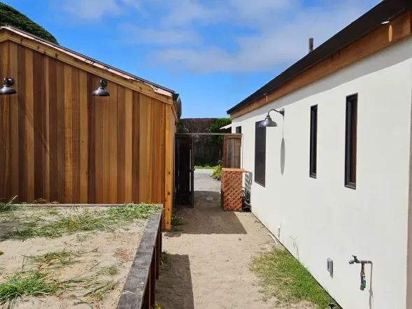 Wooden and white buildings flank a sandy path leading to a wooden gate under a blue sky.