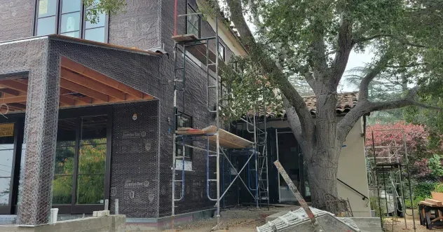 Exterior view of a house under construction with scaffolding, a large tree, and exposed siding.