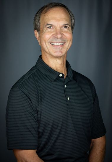 Man in black polo shirt smiles, posing against a dark gray background.