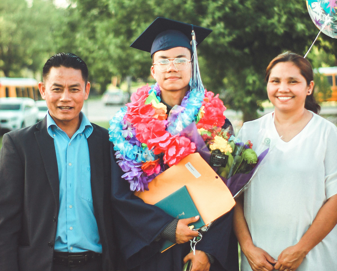 Graduate in cap and gown holding diploma, smiling with two people outdoors, wearing colorful leis.
