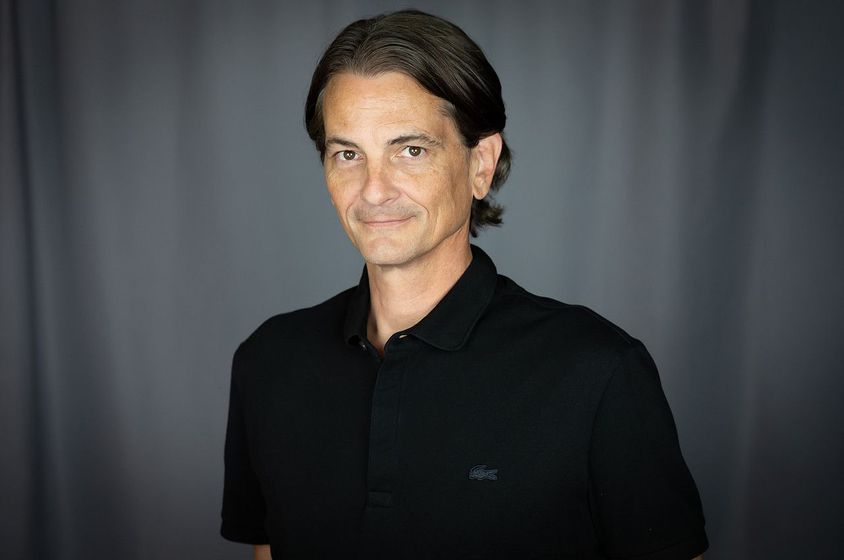 Man in a black polo shirt, smiling, against a gray backdrop.