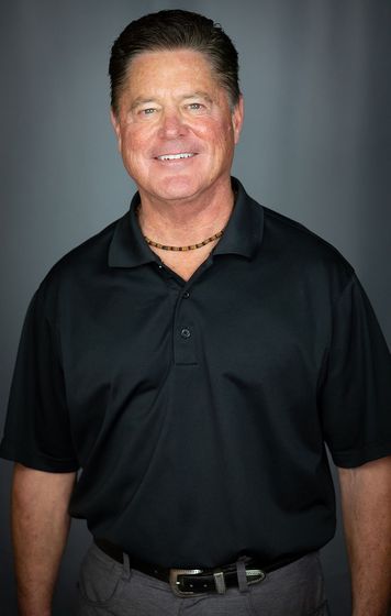 Man in black polo shirt smiles, posing against a gray background. He wears a beaded necklace.