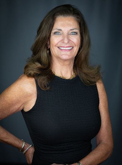 Woman in a black dress, smiling with arms at her sides, posing against a dark blue background.