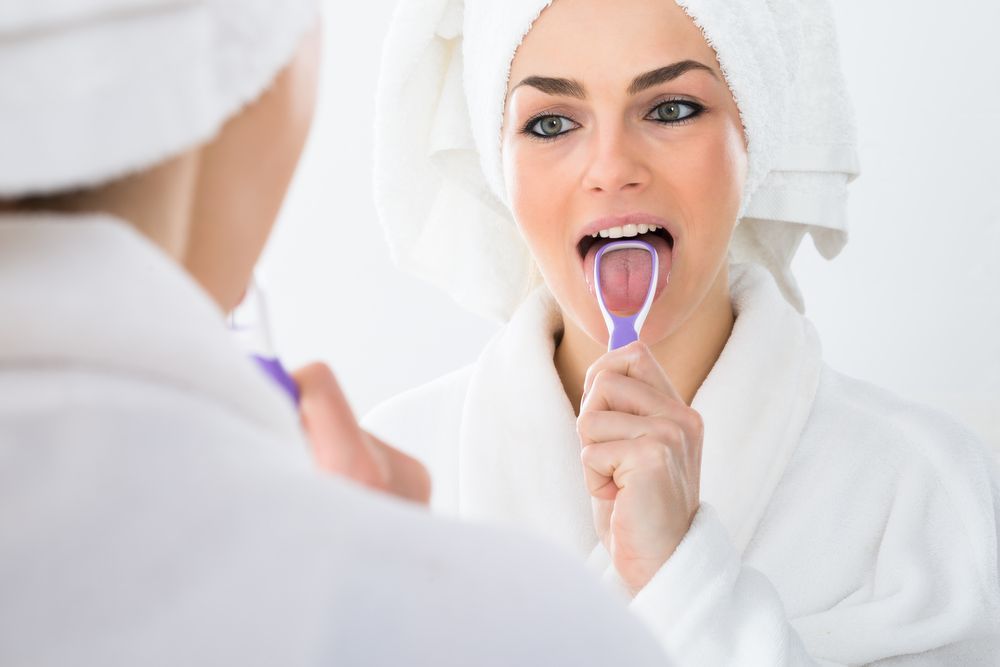 Woman using a tongue scraper in bathroom mirror. She wears a robe and has a towel on her head.