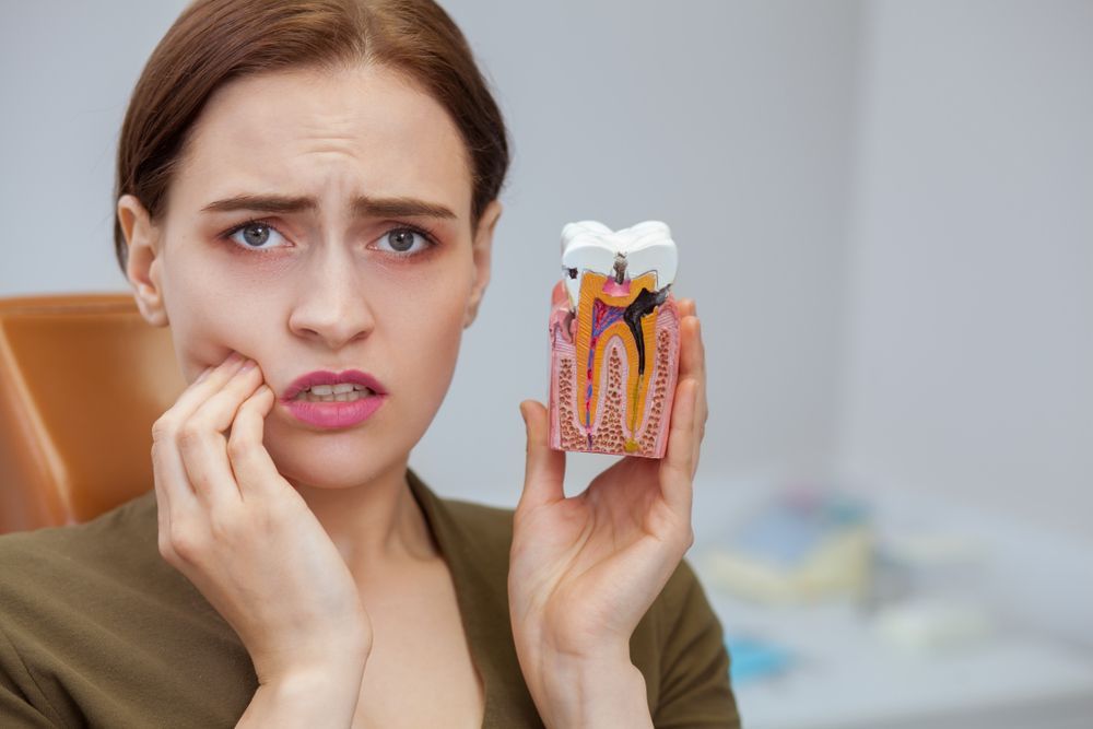 Woman holding a tooth model, grimacing and touching cheek, showing discomfort in a dental office.