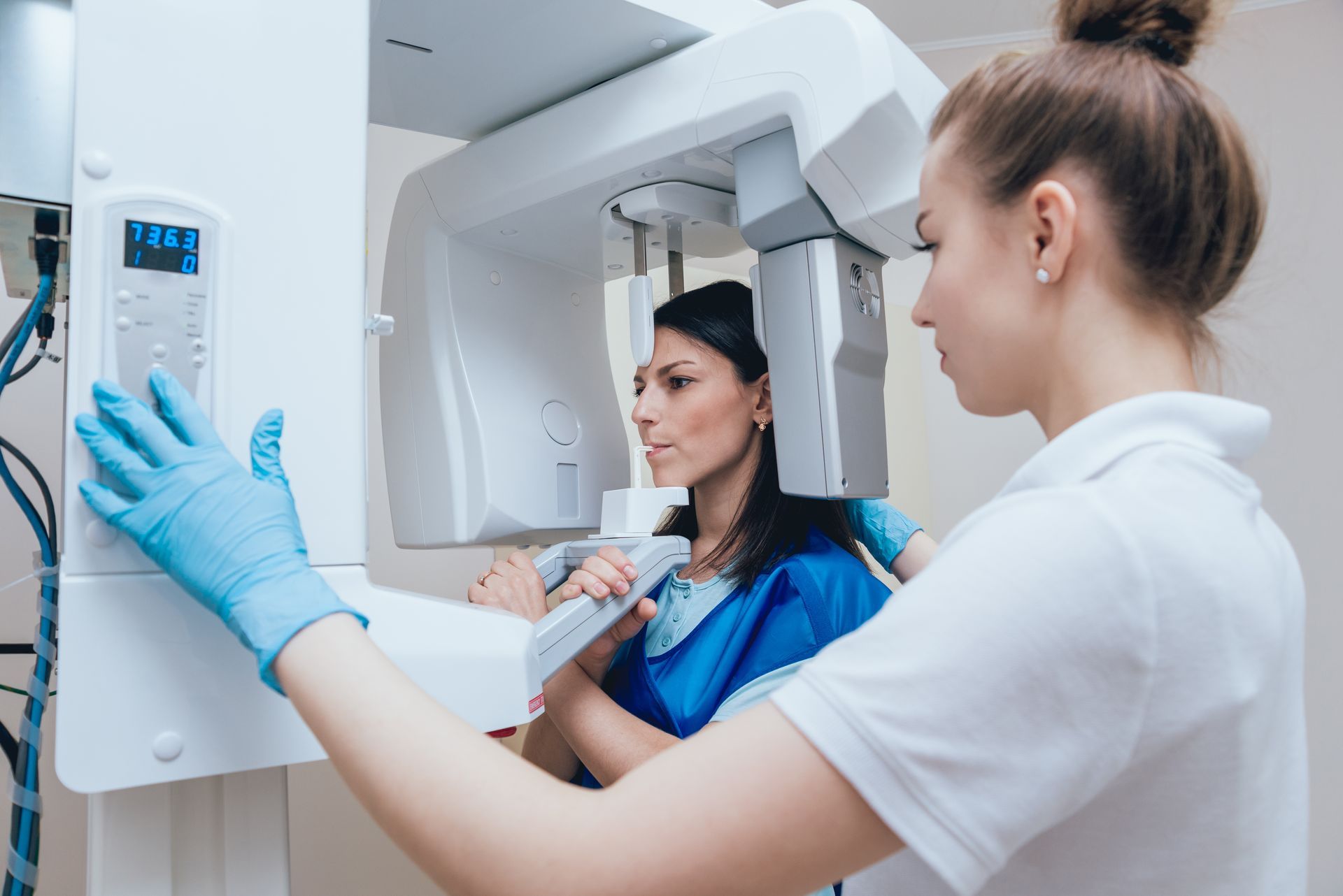 Woman receiving dental X-ray. Dental assistant in blue gloves operates machine near patient in a clinic.