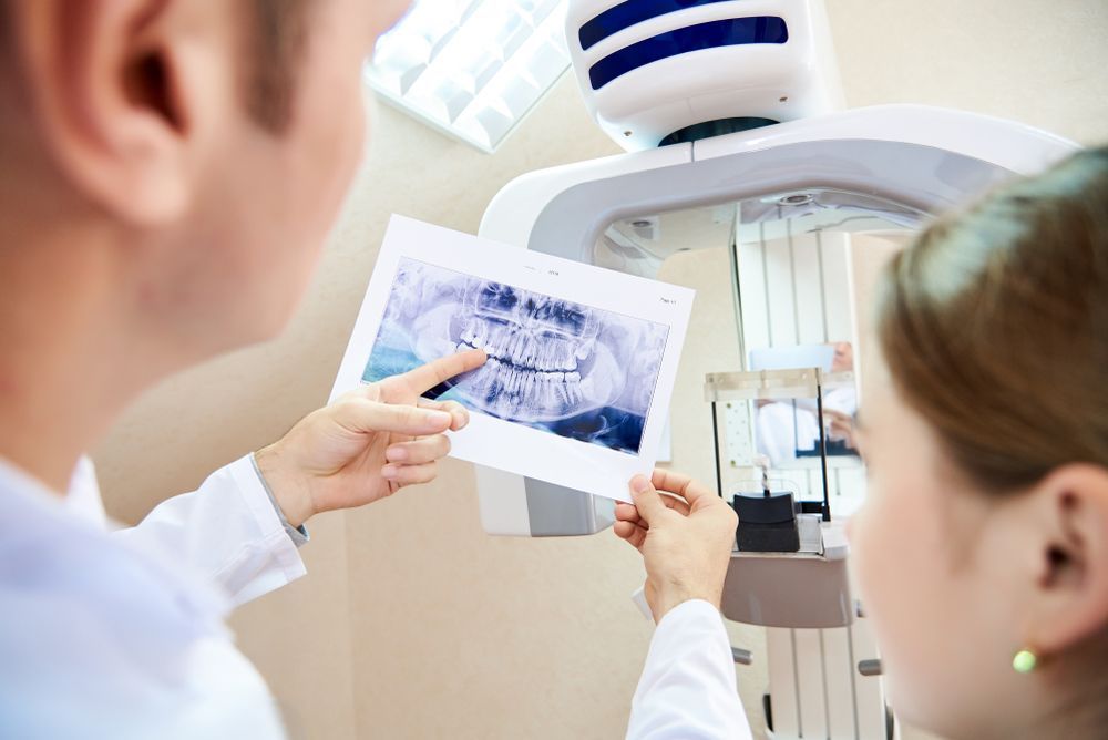 Dentist pointing at X-ray with patient; dental office setting with X-ray machine.