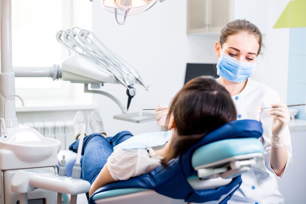Dentist wearing mask examining patient's teeth in a dental chair.