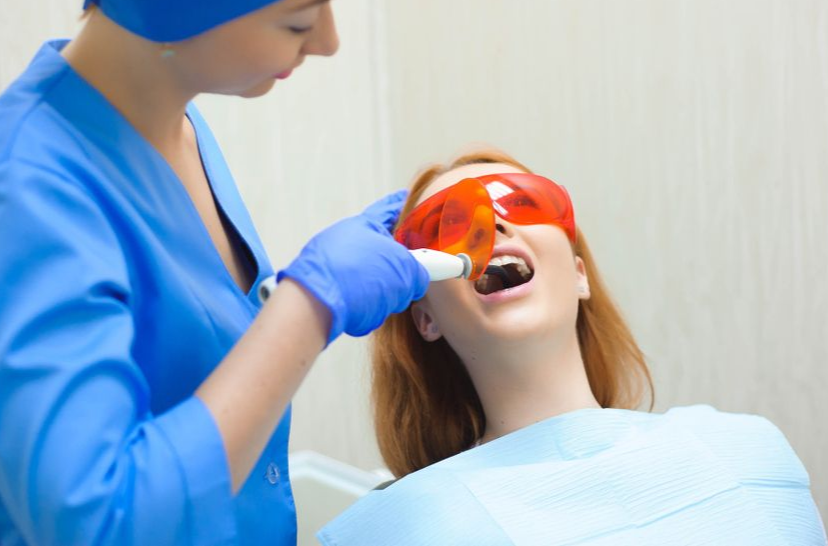 Dentist using a curing light on a patient's teeth. Patient wearing orange safety glasses, mouth open.
