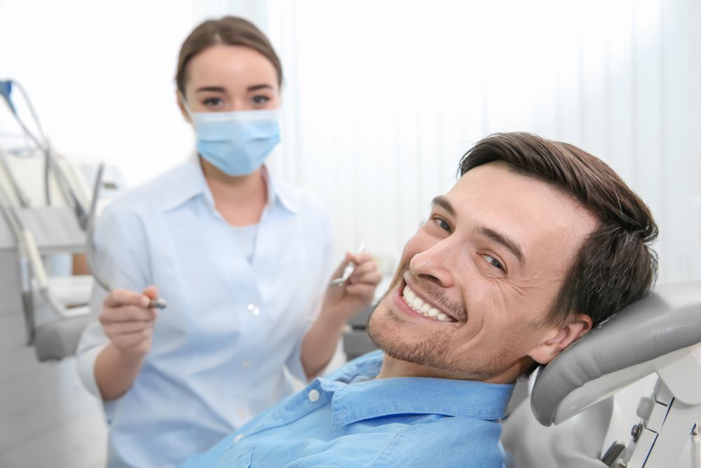 Man smiles in dentist chair; dentist in mask and uniform holds tools in the background.