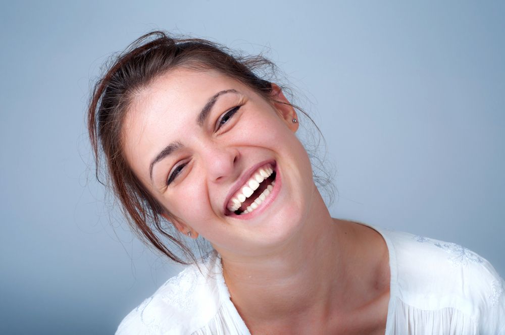 Woman with brown hair laughs, showing teeth, white shirt, light blue background.