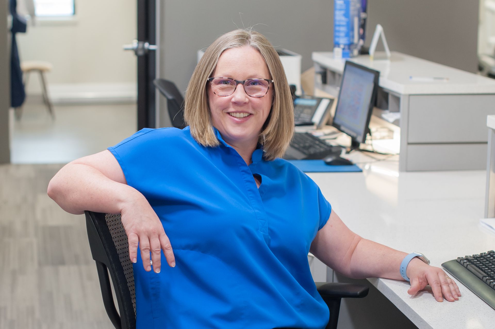 Woman wearing glasses in blue shirt sits at a desk, smiling. Modern office setting.