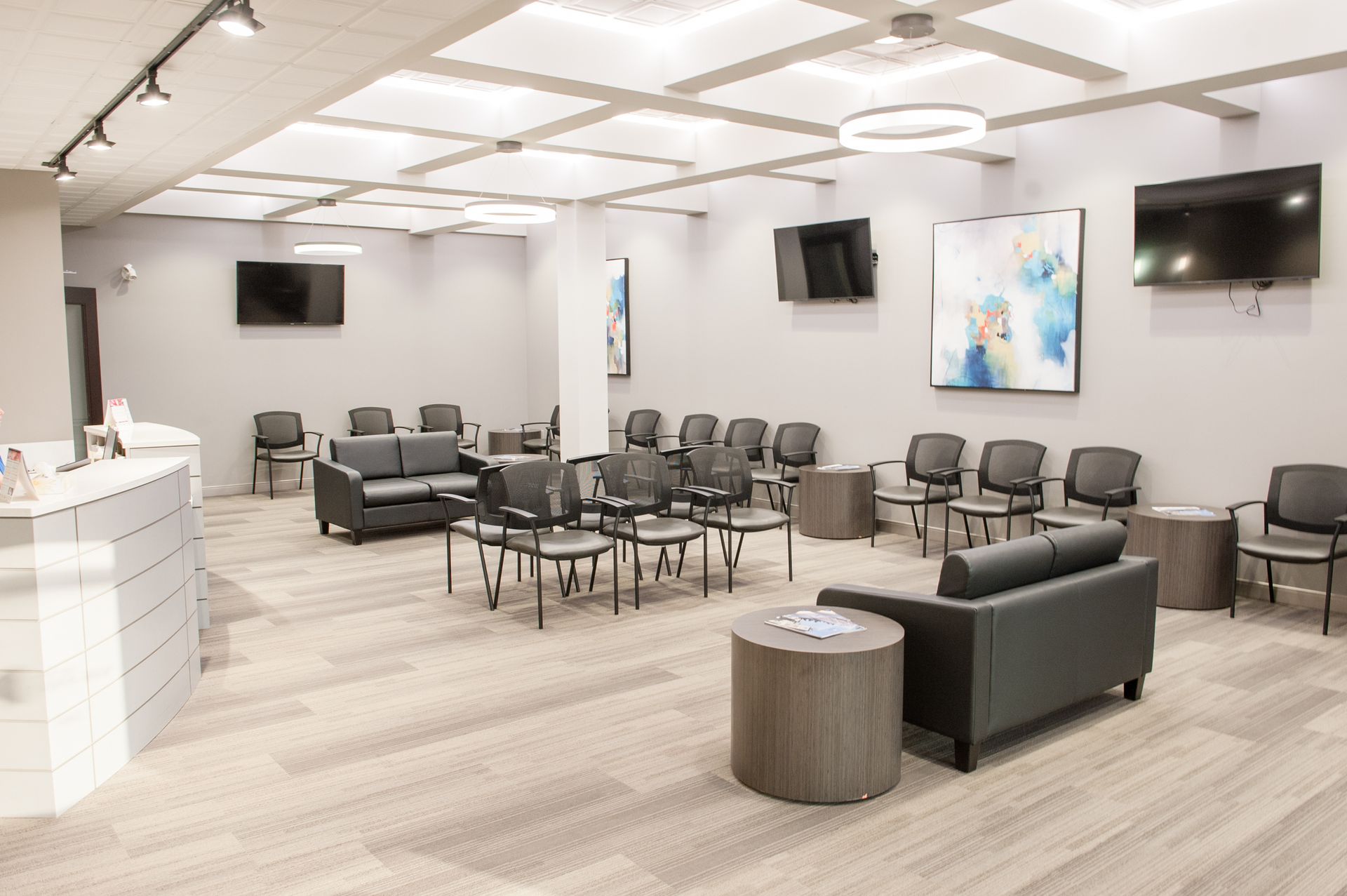 Waiting room with chairs, couches, TVs, and artwork on gray walls, with a tiled ceiling and carpeted floor.