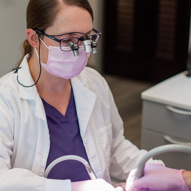Dentist examining patient's teeth with tools; wearing mask, glasses, blue gloves, and black scrubs.
